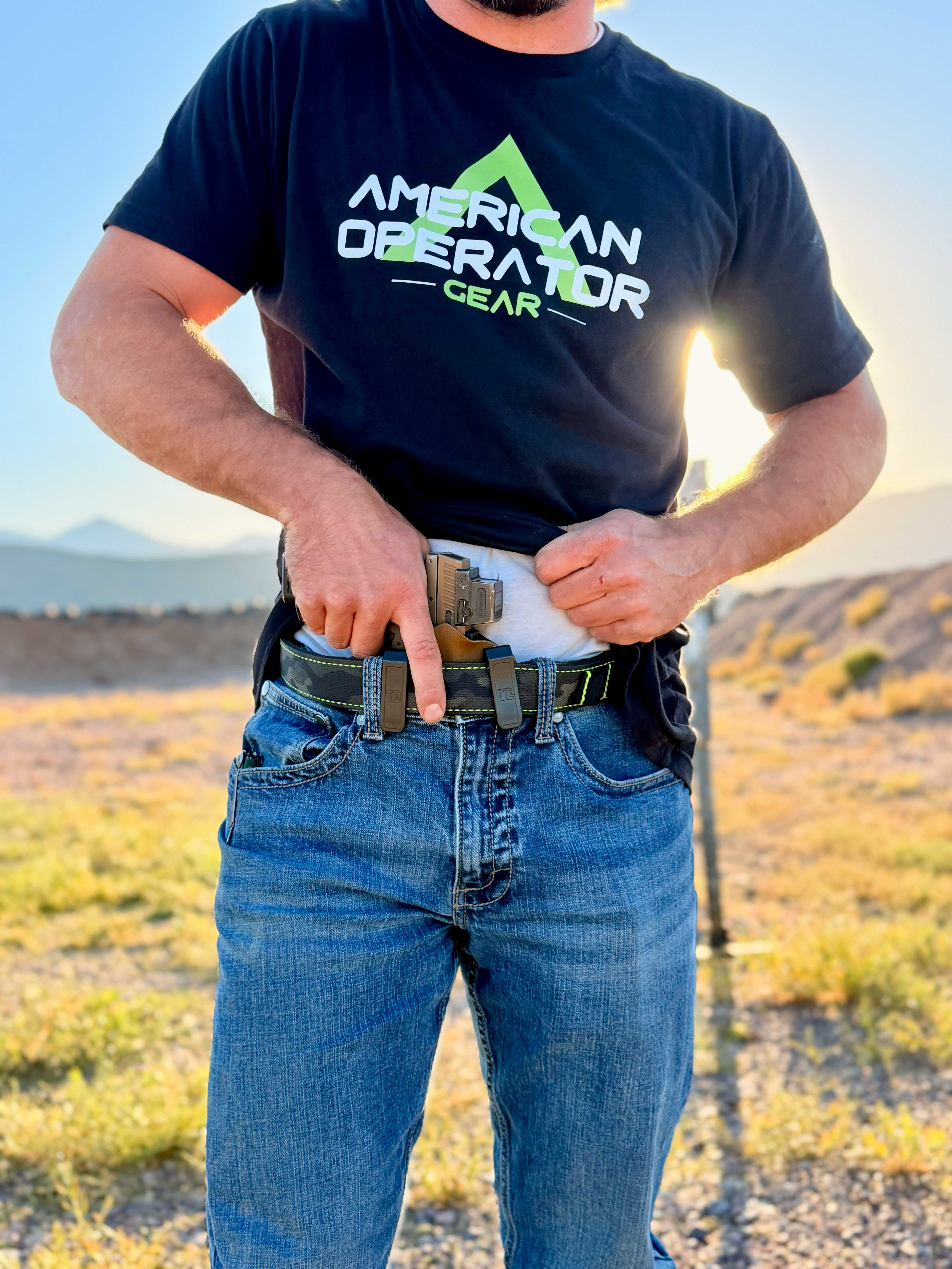 A man wearing jeans and a t-shirt adjusts his purposeful belt buckles in a scenic outdoor setting.